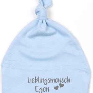 Puede incluir: Un gorro de bebé azul claro con la parte superior anudada. El gorro presenta el texto "Lieblingsmensch Egon" en gris, junto con dos símbolos de corazón grises. El gorro está hecho de tela suave.