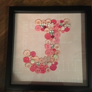 May include: A framed artwork featuring a large letter J made from pink and beige buttons. The background is a light pink with the words "baby girl" repeated in a white font.