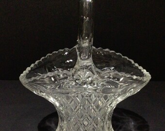 May include: A clear glass basket with a handle. The basket has a diamond pattern on the body and a scalloped edge. The handle is arched and attached to the rim. The basket is likely used for decorative purposes.