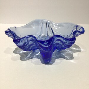 May include: A blue glass bowl with a unique, wavy design. The bowl has a light blue interior and a darker blue, ruffled edge. It is supported by three small, pointed feet. The bowl's shape is reminiscent of a flower or shell.