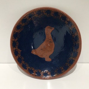 May include: A handmade ceramic bowl with a dark blue interior and a brown duck silhouette in the center. The rim is brown with a dotted pattern. This decorative bowl is perfect for serving or display.