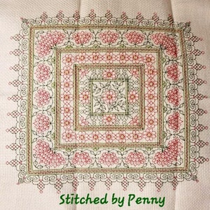 May include: A square, cross-stitched textile with a floral design in shades of pink, green, and brown on a cream background. The text "Stitched by Penny" is in green at the bottom. The design features a central square with a floral pattern.