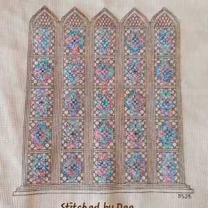 May include: A cross-stitch embroidery of a stained glass window with a repeating geometric pattern in shades of pink, blue, and green. The window is framed in brown and the text "Stitched by Dee" is at the bottom.