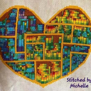 Op de afbeelding: Een hartvormig kruissteekontwerp met een geometrisch patroon in tinten blauw, groen, geel en bruin. Het ontwerp is omlijnd in goud en bevat de tekst "Stitched by Michelle" in paars.