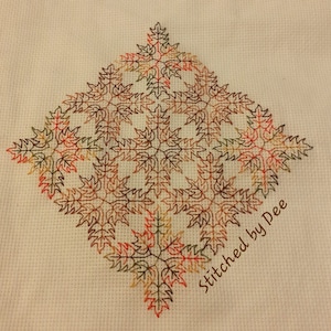 May include: A cross-stitch design featuring a geometric pattern of stylized leaves in shades of red, orange, brown, and green. The design is stitched on a white Aida cloth and includes the text "Stitched by Dee".