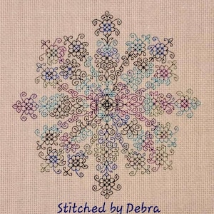 May include: A black and white cross-stitch design with a floral pattern. The design is stitched on a light pink fabric. The text "Stitched by Debra" is at the bottom of the design.