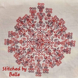 May include: A red and purple cross-stitch design with a floral pattern. The design is stitched on white Aida cloth. The text "Stitched by Belle" is in the bottom left corner of the image.