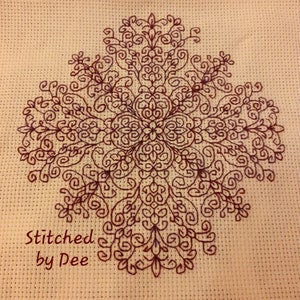 May include: A cross-stitch design featuring a symmetrical floral pattern in burgundy thread. The design is centered on a cream-colored fabric, with the text "Stitched by Dee" in the lower left corner.