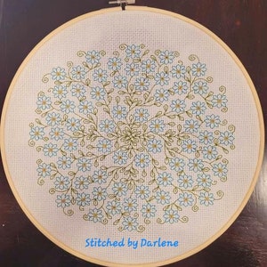 May include: A white embroidery hoop with a blue and green floral design. The design is a circle of forget-me-not flowers with green stems and leaves. The text "Stitched by Darlene" is at the bottom of the hoop.