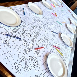 May include: A white table cover with a black and white birthday-themed design. The design includes a cake, stars, hearts, and the words "Happy Birthday". There are several white plates and crayons on the table.
