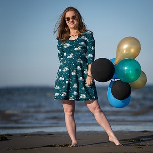 Puede incluir: Una mujer con un vestido azul verdoso con un estampado blanco y dorado de pájaros volando y nubes, sosteniendo un ramo de globos de varios colores, incluyendo negro, azul, verde y dorado. La mujer está de pie en una playa de arena con el océano de fondo.