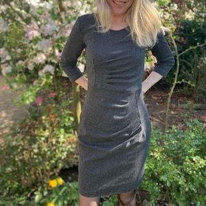 May include: A woman wearing a gray and black plaid dress with a fitted silhouette. She is standing in front of a flowering tree.