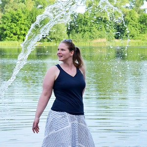 Könnte beinhalten: Eine Frau in einem marineblauen Tanktop und einem weiß-grau gemusterten Rock steht in einem See, während Wasser um sie herum spritzt. Der Hintergrund zeigt eine grüne Baumreihe und einen ruhigen See.