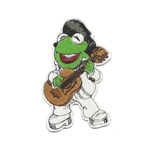 Può includere: Toppa ricamata di una rana verde che indossa un abito bianco e capelli neri, che suona una chitarra marrone.