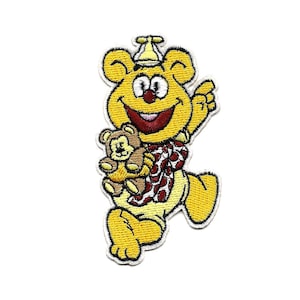 Op de afbeelding: Gele geborduurde cartoonbeer met een rood-witte geblokte sjaal en een gele hoed, die een bruine teddybeer vasthoudt.