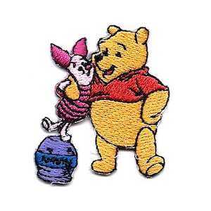 May include: Embroidered iron-on patch featuring Winnie the Pooh and Piglet. Winnie the Pooh is wearing a red shirt and Piglet is wearing a pink shirt. Piglet is standing on a purple pot with the word "Honey" on it.