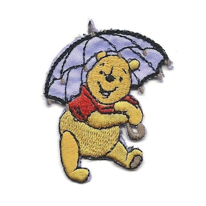 May include: Embroidered patch featuring a yellow bear wearing a red shirt and holding a light blue umbrella. The bear is smiling. The patch is designed with a stitched border and is suitable for clothing or accessories.