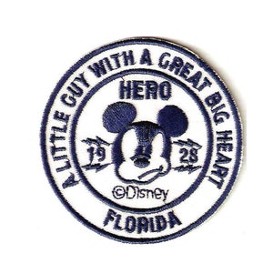 Könnte beinhalten: Ein weißer gestickter Aufnäher mit blauem Rand. Der Aufnäher zeigt ein Cartoon-Bild von Mickey Mouse mit dem Text "A Little Guy With A Great Big Heart" und "Hero" über Mickey Mouse's Kopf. Der Text "©Disney" und "Florida" befindet sich unter Mickey Mouse's Kopf.