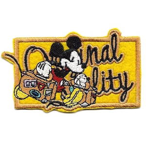 Puede incluir: Un parche bordado amarillo con Mickey Mouse sosteniendo una maleta. El texto "Original Quality" está escrito en cursiva negra.
