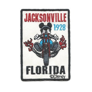 May include: A white embroidered patch with a black border. The patch features a cartoon image of Mickey Mouse riding a motorcycle with flames coming from the exhaust. The text "JACKSONVILLE 1928 FLORIDA" is embroidered in red and black. The text "©Disney" is embroidered in black at the bottom of the patch.