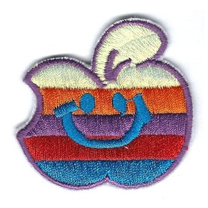 Pode incluir: Um remendo bordado colorido em forma de maçã com um rosto sorridente. A maçã é listrada de laranja, vermelho, roxo, azul e branco.