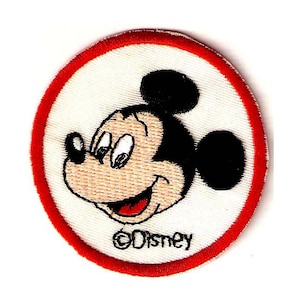 Puede incluir: Un parche blanco bordado con un borde rojo que presenta la cara de Mickey Mouse. El parche tiene el texto "©Disney" en la parte inferior.