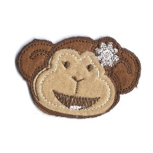 May include: A felt monkey face applique in shades of brown and tan, with a stitched smile and a silver flower accent. The applique is designed for crafts and sewing projects.