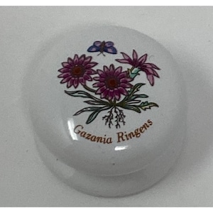 May include: White ceramic lid with a floral design featuring purple Gazania Ringens flowers and a blue butterfly. The flowers have dark purple centers and green leaves. The text "Gazania Ringens" is printed in brown.