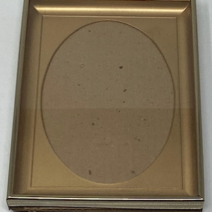 May include: A rectangular gold-colored picture frame with an oval opening. The frame has a simple, elegant design with a thin border and a neutral-colored backing. The frame is designed to hold a photograph or artwork.