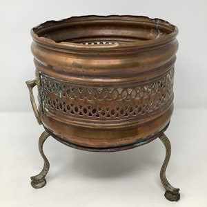 May include: A copper plant stand with an intricate, openwork design and three legs. The stand is made of polished copper and has a rustic, antique look.