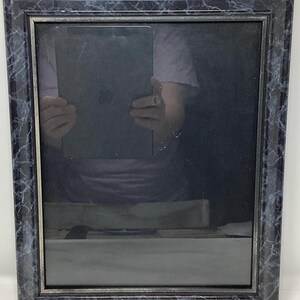 May include: A rectangular picture frame with a dark blue marble pattern. The frame surrounds a reflective surface, with a hint of a person holding an object. The frame's design adds a touch of elegance.