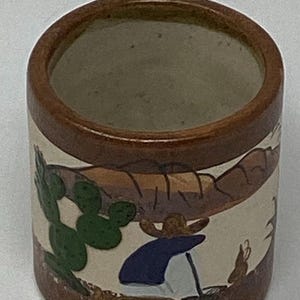 May include: A ceramic mug with a brown rim, handle, and base. The mug features a hand-painted scene with a cactus, a person in a sombrero, and a desert landscape in shades of brown, green, and blue.