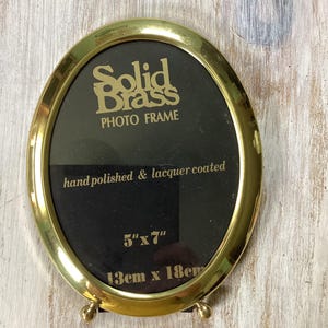 May include: Oval brass photo frame with the text "Solid Brass PHOTO FRAME". The frame is hand polished and lacquer coated, and measures 5" x 7". It has a black interior and small ball feet.