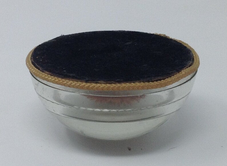 May include: A silver-coloured, round, metal container with a black velvet lining and a gold trim around the top.