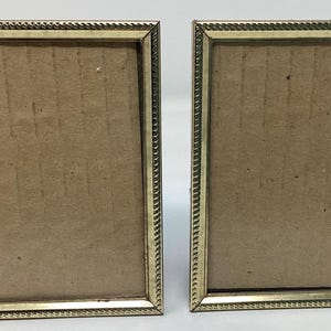 May include: Two gold-colored picture frames with a simple, ornate design. The frames are empty and ready to display photos.
