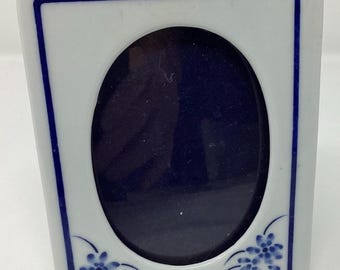 May include: White porcelain picture frame with a blue and white floral design. The frame has an oval opening for a photo.