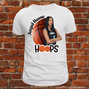 Puede incluir: Camiseta blanca con un gráfico de baloncesto y el texto "Angel Reese HOOPS". La pelota de baloncesto es naranja y negra y tiene una cara de mujer en ella.