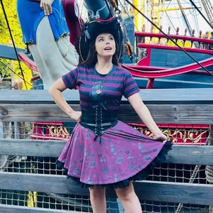 Puede incluir: Una mujer con una camisa a rayas blancas y negras, un corsé negro y una falda morada con estampado blanco y negro. Lleva un sombrero de pirata negro y está de pie frente a un barco.