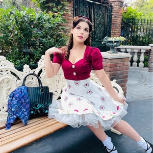 Puede incluir: Una mujer con un top burdeos con botones blancos y una falda blanca con estampado de margaritas. Está sentada en un banco blanco, con un bolso negro y una bufanda azul. Lleva zapatos negros y calcetines blancos.