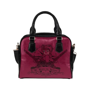 May include: A burgundy handbag with a black strap and handles. The front of the bag features a black and white illustration of a woman with a red bandana and two pistols. The text "We Wants the Redhead" is printed on a banner below the illustration.
