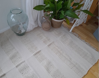 May include: A cream-colored rug with a geometric pattern of diamonds. The rug has a fringe border and is laid out on a wooden floor. A potted plant with white flowers is in the background.