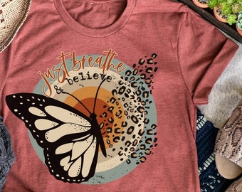 Könnte beinhalten: Ein rostfarbenes T-Shirt mit einem Schmetterlingsmotiv und dem Text "Just Breathe & Believe" im Retro-Stil.