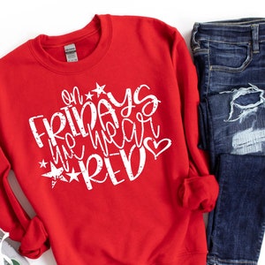 K&ouml;nnte beinhalten: Roter Pullover mit wei&szlig;em Text, der "On Fridays we wear red" mit Sternen und einem Herz sagt. Der Pullover liegt auf einer wei&szlig;en Oberfl&auml;che mit einem Paar grauer Turnschuhe und einer blauen Jeans.