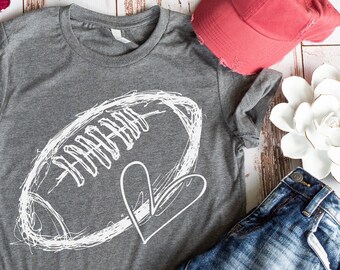 May include: Gray t-shirt with a white outline of a football and a heart. The shirt is laid out on a wooden surface with a red and white striped sweater, a pair of red and white striped shoes, a pair of blue jeans, a red baseball cap, and a white flower.
