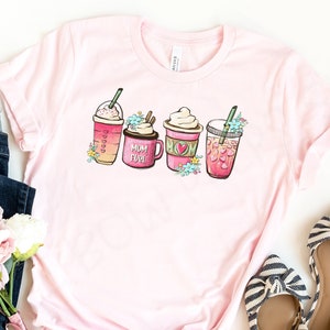 Op de afbeelding: Een roze T-shirt met een afbeelding van vier verschillende koffiedrankjes. De drankjes zijn versierd met harten en bloemen. De tekst "Mom Fuel" staat op een van de drankjes.