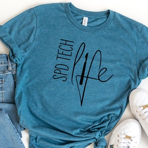 Könnte beinhalten: Ein blaues T-Shirt mit dem Text "SPD TECH LIFE" und einem stilisierten Bild einer medizinischen Spritze. Das Shirt wird mit einer blauen Jeans und weißen Turnschuhen kombiniert.