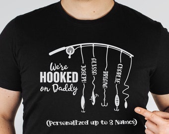 May include: Black t-shirt with a white graphic of a fishing rod with hooks and the text "We're HOOKED on Daddy (Personalized up to 8 Names)". The hooks have names written on them: Alissa, Joseph, Charlie, Briana.