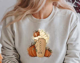 Può includere: Una felpa bianca con una grafica di un latte alla zucca speziato con panna montata e zucche. Il testo "PSL" è sulla tazza.