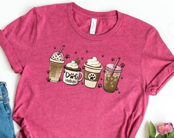 以下が含まれることがあります： 白い背景に置かれたピンクのTシャツ。Tシャツには、ホイップクリームとストローが付いたコーヒーカップ4つのグラフィックがプリントされている。カップの1つには「Dog Mom」の文字が書かれている。Tシャツの隣には、白いスニーカーと青いジーンズが置かれている。