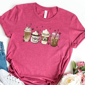Pode incluir: Uma camiseta rosa com um gr&aacute;fico de quatro x&iacute;caras de caf&eacute; com chantilly e um canudo. O texto em uma das x&iacute;caras diz "Dog Mom". A camiseta est&aacute; colocada sobre uma superf&iacute;cie branca com um par de t&ecirc;nis brancos e uma cal&ccedil;a jeans azul.